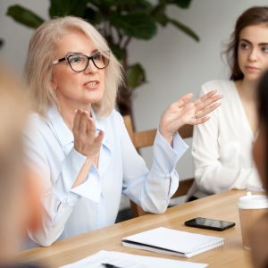 Attractive aged businesswoman, teacher or mentor coach speaking to young people, senior woman in glasses teaching audience at training seminar, female business leader speaker talking at meeting
