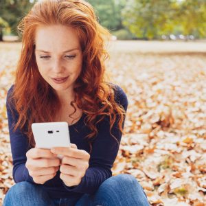 Attractive young woman sitting amongst fallen autumn leaves in a park reading an sms or text message on her mobile phone with copy space